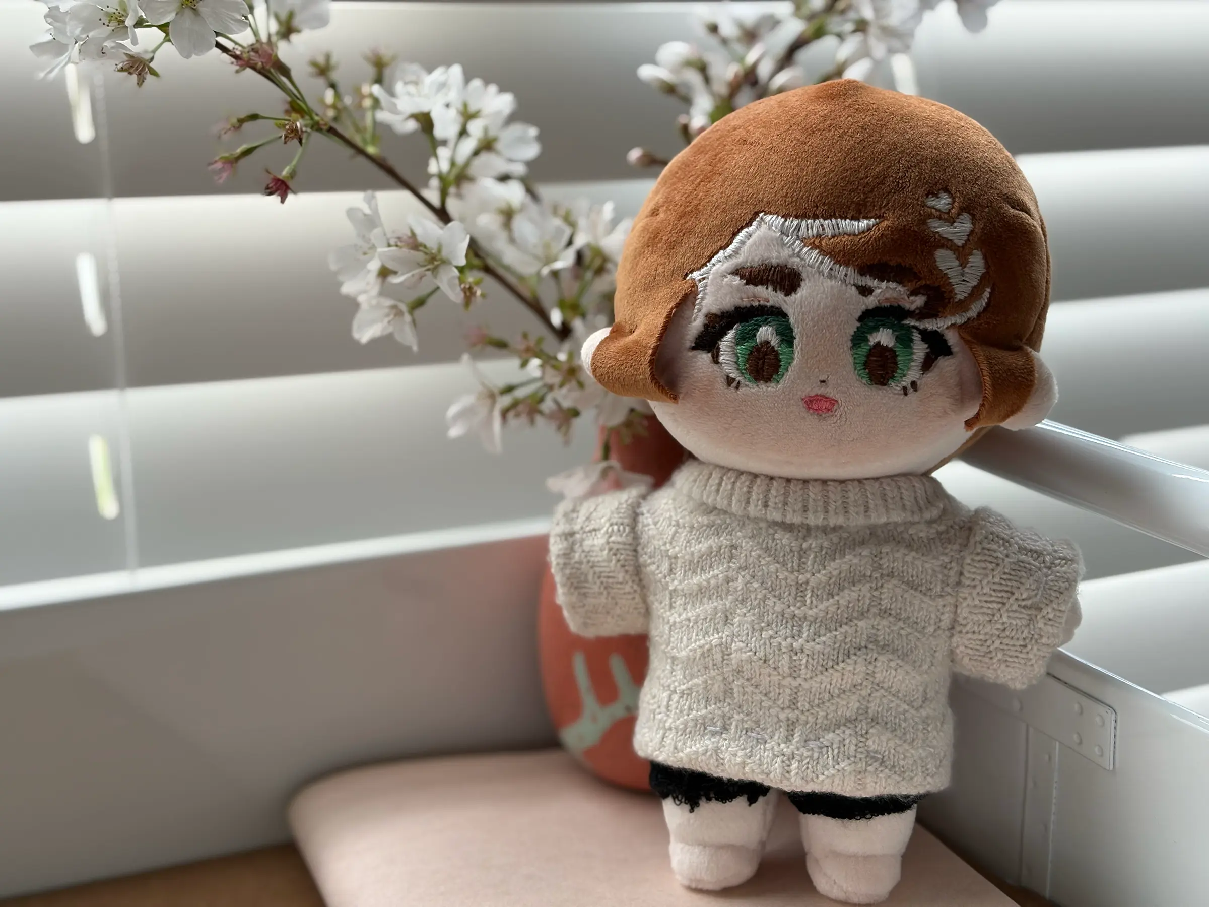 Handmade doll with brown hair and green eyes in a cream cable-knit sweater, posed beside white cherry blossoms.